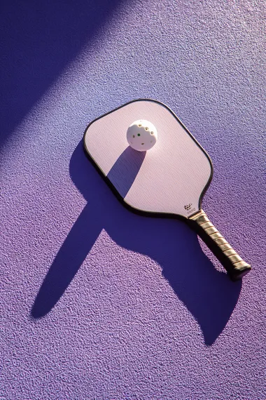 trendy pickleball paddle with ball on textured lavender surface, high angle summer sunlight casting bold shadow, minimal composition, no text