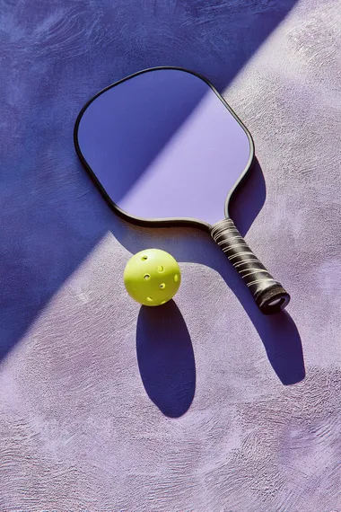 trendy pickleball paddle with ball on textured lavender surface, high angle summer sunlight casting bold shadow, minimal composition, no text