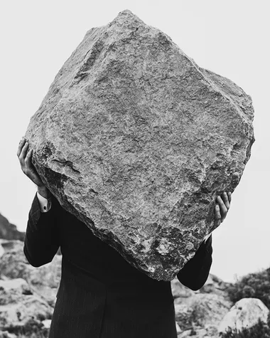 black and white surreal photo of a man in a suit shown from the waist up, carrying a large rough stone on his back, slightly hunched forward, close-up framing with clear detail on the fabric, skin tension, and texture of the stone, soft background blur with minimal horizon, poetic editorial surrealism, cinematic shadow, high-grain analog film texture, modern minimalist composition   --raw rofile ljxxpnm