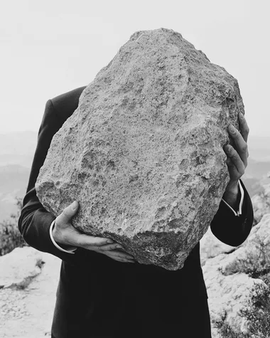 black and white surreal photo of a man in a suit shown from the waist up, carrying a large rough stone on his back, slightly hunched forward, close-up framing with clear detail on the fabric, skin tension, and texture of the stone, soft background blur with minimal horizon, poetic editorial surrealism, cinematic shadow, high-grain analog film texture, modern minimalist composition   --raw rofile ljxxpnm