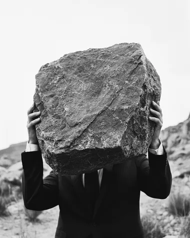 black and white surreal photo of a man in a suit shown from the waist up, carrying a large rough stone on his back, slightly hunched forward, close-up framing with clear detail on the fabric, skin tension, and texture of the stone, soft background blur with minimal horizon, poetic editorial surrealism, cinematic shadow, high-grain analog film texture, modern minimalist composition   --raw rofile ljxxpnm