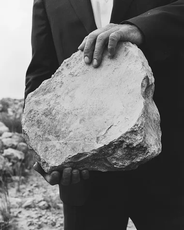 black and white surreal photo of a man in a suit shown from the waist up, carrying a large rough stone on his back, slightly hunched forward, close-up framing with clear detail on the fabric, skin tension, and texture of the stone, soft background blur with minimal horizon, poetic editorial surrealism, cinematic shadow, high-grain analog film texture, modern minimalist composition   --raw rofile ljxxpnm