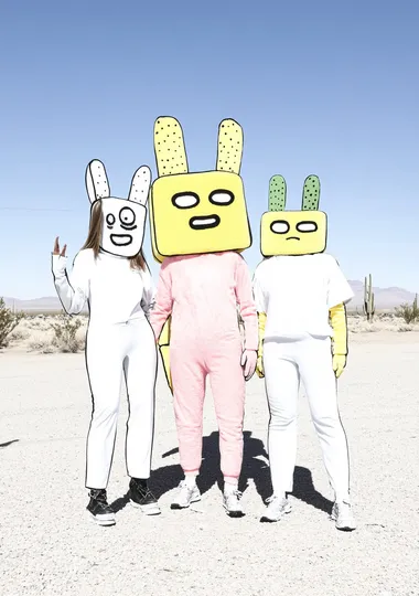 three female humanoids wearing oversized plush pink and yellow robot heads with cactus arms. On their shoulders is a huge art installation made from giant speakers. The background is a desert landscape.   1ug8ngh