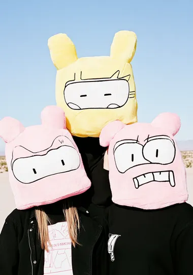 three female humanoids wearing oversized plush pink and yellow robot heads with cactus arms. On their shoulders is a huge art installation made from giant speakers. The background is a desert landscape.   1ug8ngh