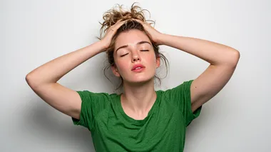 A candid portrait of an overheated young white woman in a green t-shirt, using her hands to fan air toward her face in an attempt to cool down, her hair slightly messy and sticking to her forehead, standing in a professional photo studio with a clean seamless white background, artificial studio lighting mimicking harsh midday sunlight, shot with a Nikon Z9, 85mm f/1.2 lens, realistic skin tones, warm highlights and natural heat-induced details