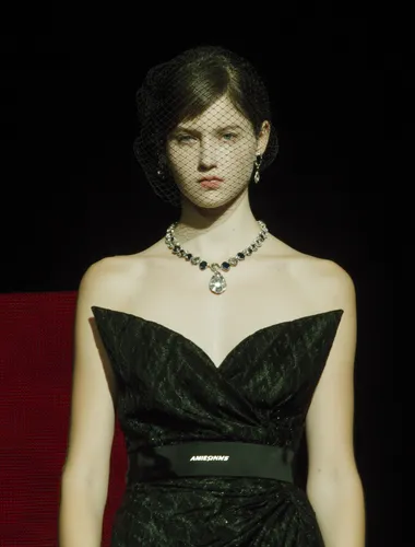 A model walks down the runway at Fashion Week wearing a black ribbon tied into a black and dark green bow, a black veil covering her face, an exquisite black dress similar to the "Amagi bag" style, and a silver teardrop-shaped gemstone around her neck. Around her neck she wears a gray-black diamond necklace with strings of tiny dark blue gemstones. The photo was taken from the front, capturing the model's upper body and head, in a style reminiscent of Alexander McQueen's "Red Book" show at Paris Fashion Week. The photo has a black background and is in HD resolution.   --raw rofile jg6bzz9