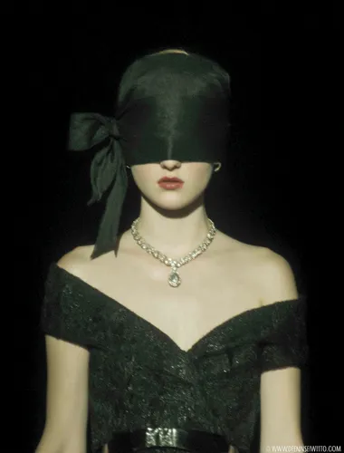 A model walks down the runway at Fashion Week wearing a black ribbon tied into a black and dark green bow, a black veil covering her face, an exquisite black dress similar to the "Amagi bag" style, and a silver teardrop-shaped gemstone around her neck. Around her neck she wears a gray-black diamond necklace with strings of tiny dark blue gemstones. The photo was taken from the front, capturing the model's upper body and head, in a style reminiscent of Alexander McQueen's "Red Book" show at Paris Fashion Week. The photo has a black background and is in HD resolution.   --raw rofile jg6bzz9