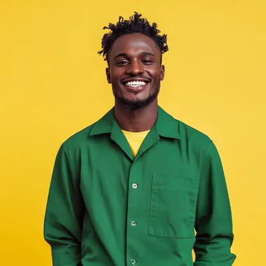 Studio shot of a 30 yr african male on a solid yellow background smiling and wearing a collared green long sleeve shirt and pants.
