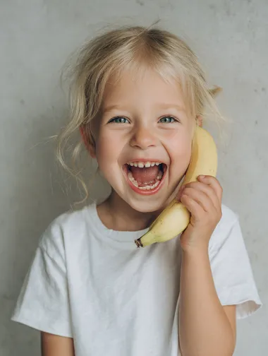 a professional photo, ultra close-up of a cute little blonde girl, white t-shirt, laughing and pretending to talk on the phone but holding an bannana like phone, shallow depth of field, studio lighting, natural joyful expression, high detail, modern interior soft focus background, realistic skin texture, editorial style  --raw