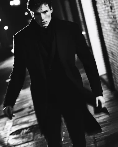 A Saint Laurent-clad luxury realtor strides through a dimly lit urban setting at dusk. His slim-cut black wool suit and silk scarf catch the movement, blurred slightly as he turns toward the camera. The lighting is dramatic, reminiscent of film noir, with long shadows stretching behind him. Shot in black and white with soft grain, evoking a timeless, rebellious elegance.