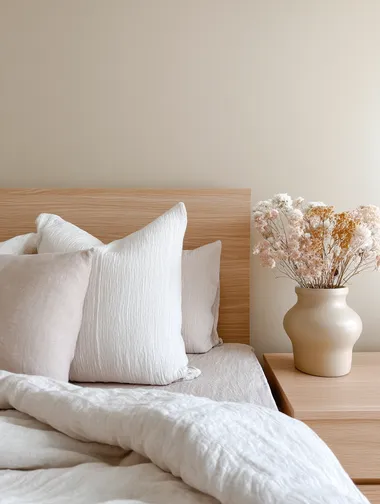 Softly styled bed corner with crinkled linen sheets, light wood headboard, calming beige wall, and dried flowers in a ceramic pot  rofile 22tuiyk
