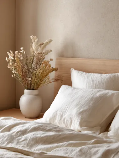 Softly styled bed corner with crinkled linen sheets, light wood headboard, calming beige wall, and dried flowers in a ceramic pot  rofile 22tuiyk