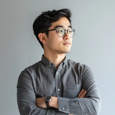 an asian male with glasses he is facing to the side and has his arms crossed, he is wearing a long sleeved button down shirt, he has a determined look on his face