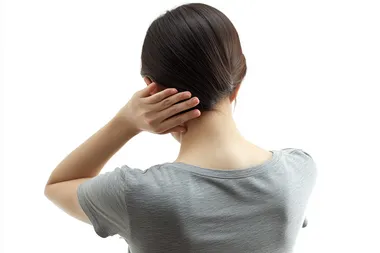Photo of a Korean woman standing with her back to the camera, wearing a casual grey t-shirt. Her left hand gently touching the back of her neck, expressing mild discomfort, with natural posture and realistic hand positioning. Soft lighting, white background, no dramatic expression, photorealistic