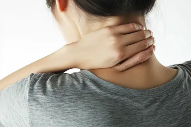 Photo of a Korean woman standing with her back to the camera, wearing a casual grey t-shirt. Her left hand gently touching the back of her neck, expressing mild discomfort, with natural posture and realistic hand positioning. Soft lighting, white background, no dramatic expression, photorealistic