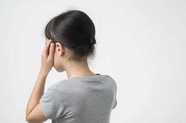 Photo of a Korean woman standing with her back to the camera, wearing a casual grey t-shirt. Her left hand gently touching the back of her neck, expressing mild discomfort, with natural posture and realistic hand positioning. Soft lighting, white background, no dramatic expression, photorealistic