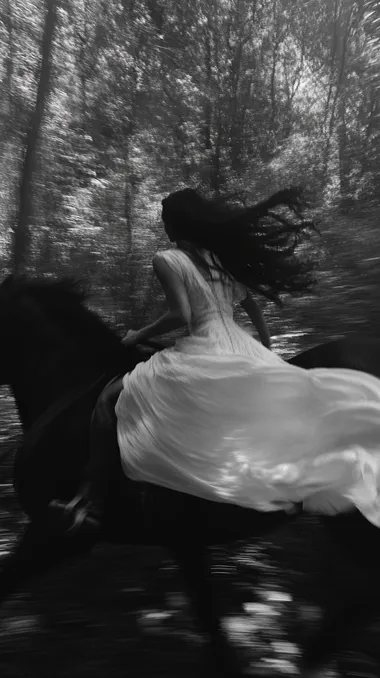A blurred black and white image of a woman in a flowing white dress riding a horse at full speed through a dense, shadowy forest. Her hair and fabric trail behind her with intense motion. The scene is shot like a frame from a forgotten 1970s art film high contrast, soft focus, and visible heavy 35mm film grain. The lighting is natural and directional, with flickers of light breaking through the trees, creating a ghostly atmosphere. The image feels emotional and raw, evoking urgency, solitude, and mythological transformation. A contemporary vision of the heros call powerful, cinematic, and dreamlike.   rofile 5shn6qo