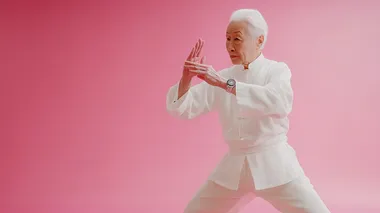 Studio style, shot in a studio setting. In the middle scene, a Chinese elderly person is practicing Tai Chi with a smartwatch on their wrist, elegant movements in white clothing, silver white hair, and a hint of grass under their feet. Filmed in a studio setting, the scene features strong contrast, high saturation, numerous details, exquisite smoothness, high definition, Rembrandt lighting, movie quality, large wide-angle, panoramic, and deep depth of field. There's nothing around them, the entire background and ground are pink, they were filmed in the studio,