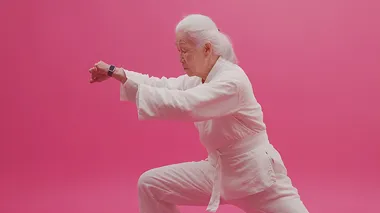 Studio style, shot in a studio setting. In the middle scene, a Chinese elderly person is practicing Tai Chi with a smartwatch on their wrist, elegant movements in white clothing, silver white hair, and a hint of grass under their feet. Filmed in a studio setting, the scene features strong contrast, high saturation, numerous details, exquisite smoothness, high definition, Rembrandt lighting, movie quality, large wide-angle, panoramic, and deep depth of field. There's nothing around them, the entire background and ground are pink, they were filmed in the studio,