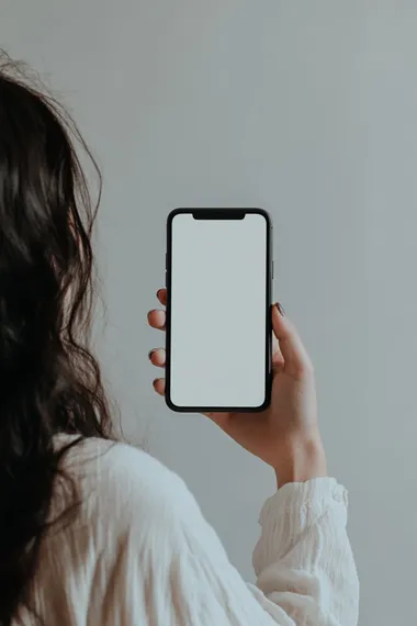 A woman holding an iPhone with a white screen, in the mockup style. The background is pure white and the focus of attention is on her hand holding up the phone. She has dark hair and appears to be in a profile view. Her fingers appear poised over the button that blends into the black frame of the phone, which she holds against a clean white backdrop. There is no visible text or other elements inside the smartphone display, highlighting its blank space for custom content or advertising copy. It is an ultra-realistic photograph captured at high resolution 32k.