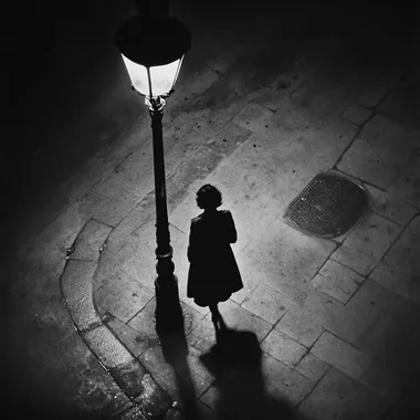 a black and white street photography style of a shadow of a woman walking alone in the street underneath a street lamp