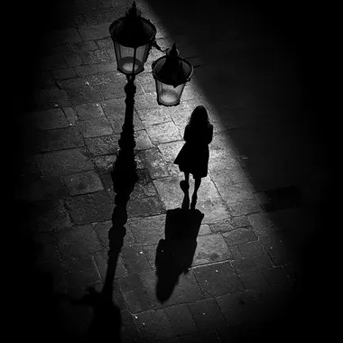 a black and white street photography style of a shadow of a woman walking alone in the street underneath a street lamp