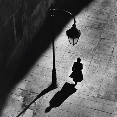 a black and white street photography style of a shadow of a woman walking alone in the street underneath a street lamp
