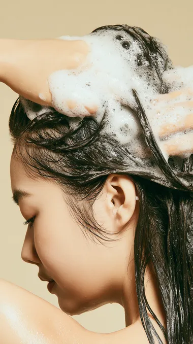 a k-pop woman shampooing hair, wet long hair falling downward with gravity, both hands massaging scalp with rich dense foam, creamy white bubbles covering hair, side composition with woman on the left, empty space on the right for text placement, partial face visible, minimal studio background in soft beige, hyper-realistic hair texture, premium shampoo commercial aesthetic, , 4k resolution, high-end Korean cosmetic brand mood, professional product photography  --s 50