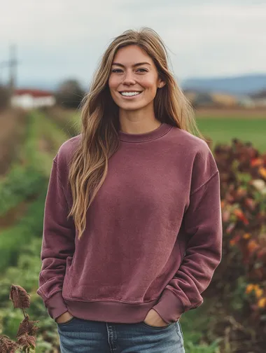 woman wearing an acid washed mauve colored crewneck sweatshirt. woman is smiling. woman is facing the camera. apparel mockup style. focus on the quality of the crewneck. background is a country scene.
