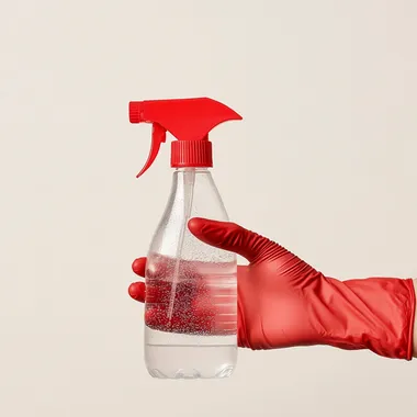 highly detailed product photo of a hand with a red latex glove on holding a clear spray bottle with a red nozzle. The bottle is half filled with clear still water. the background is a clean cream white gradient with bright natural light. photorealistic, 4K.