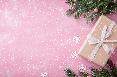 Christmas-themed pink background with white snowflakes, Christmas tree branches, and a gift box on a pastel pink tabletop. Flat lay style. Bright colors, high resolution. The concept of holiday celebration and shopping in the festive season. Stock photo with 2/3 space for text.  --s 50  sqq5bir