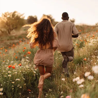photorealistic DSLR still photo - young attractive Black couple running through a wildflower meadow of poppies, daisies and lupines, captured from behind, mid-stride; woman with long loose curly hair flowing, wearing a sundress; man wearing light taupe linen shirt and stone chinos; Canon EOS R5, 135 mm f/2 lens, shutter 1/2000 s, ISO 100, warm golden-hour back-rim light, cinematic depth of field, subtle dust motes, ultrarealistic texture, 8-k detail  watermark --raw