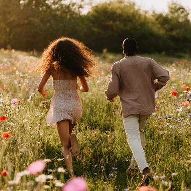 photorealistic DSLR still photo - young attractive Black couple running through a wildflower meadow of poppies, daisies and lupines, captured from behind, mid-stride; woman with long loose curly hair flowing, wearing a sundress; man wearing light taupe linen shirt and stone chinos; Canon EOS R5, 135 mm f/2 lens, shutter 1/2000 s, ISO 100, warm golden-hour back-rim light, cinematic depth of field, subtle dust motes, ultrarealistic texture, 8-k detail  watermark --raw