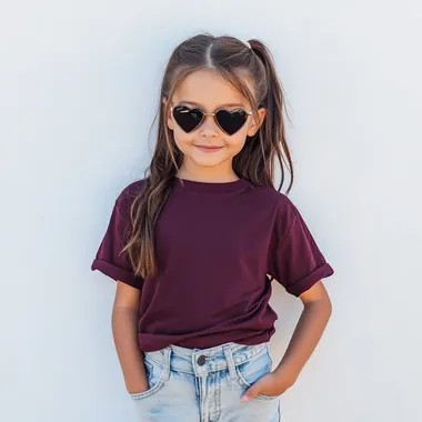 adorable 9 year old girl, wearing an oversized maroon color comfort colors T-shirt, the t-shirt has cuffed sleeves, high waisted light denim jeans, facing forward full view of tshirt, she is also wearing black heart shaped sunglasses outside in natural lighting front of a white wall in summer, trendy, posing, smiling --raw